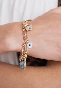 Gouden metalen armband met een vlinderbedel, een blauwe oogkraaltje en kleine decoratieve accenten. Kettinglinks met verschillende texturen.