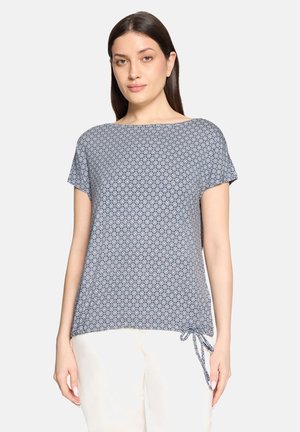 Frau trägt ein kurzärmliges, blau gemustertes Oberteil mit rundem Ausschnitt und einer seitlichen Bindung, kombiniert mit weißen Hosen, steht vor einem schlichten Hintergrund.