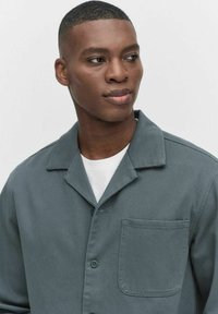 Grijs doorgestikte shirt van textiel met een voorzak en knoopsluiting. Daaronder zit een wit t-shirt.