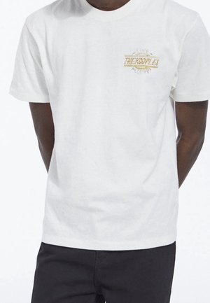 Man draagt een wit T-shirt met korte mouwen met een klein geel logo "The Kooples Records" op de linkerbovenborst, gecombineerd met een zwarte broek.