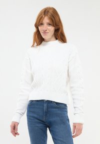 Biały sweter z włóczki z wysokim kołnierzem i wzorem w warkocze, z prążkowanymi mankietami i długimi rękawami, w połączeniu z niebieskimi dżinsami.