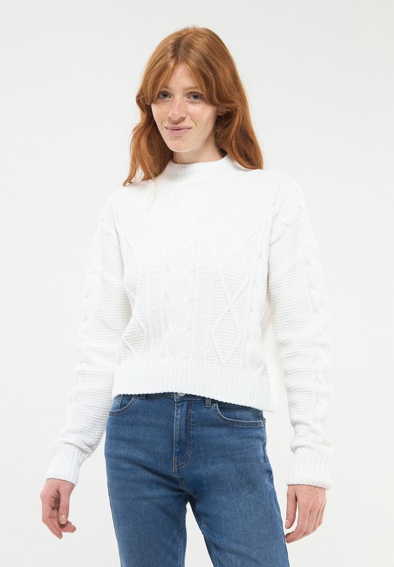 Biały sweter z włóczki z wysokim kołnierzem i wzorem w warkocze, z prążkowanymi mankietami i długimi rękawami, w połączeniu z niebieskimi dżinsami.