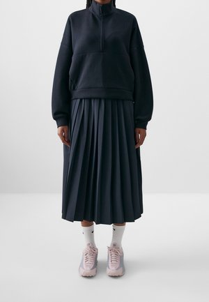 Person iført en sort kvart-lynlås sweatshirt, sort plisseret midinederdel, hvide Nike crew-sokker og pinke sportssko, stående mod en hvid baggrund.