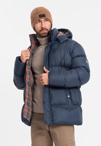 Giacca a vento blu navy con fodera a quadri, cappuccio e tasche laterali. Il modello indossa un pullover beige e un berretto di lana marrone.