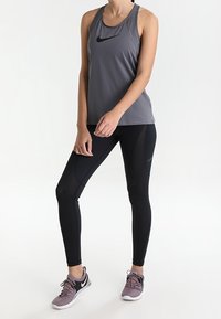 Šedý sportovní tílko s černým logem, spárováno s černými legínami s síťovinovými detaily. Nosené se šedými sportovními botami.