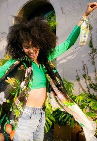 Șal negru cu flori, cu modele colorate, aruncat peste un top verde deschis și pantaloni scurți din blug albastru deschis, amplasat pe un fundal de frunziș verde.