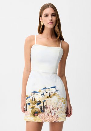 Kobieta w białej sukience na cienkich ramiączkach z namalowanym krajobrazem przedstawiającym domy z niebieskimi dachami i drzewa na spodnicy.