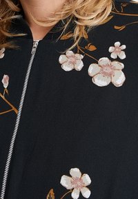 Giacca nera con ricami floreali in bianco e rosa chiaro. Presenta una cerniera argentata e un tessuto strutturato e liscio.