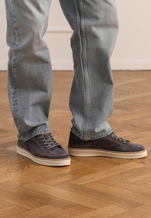 Person wearing light blue denim jeans and dark gray suede sneakers standing on a wooden parquet floor indoors.