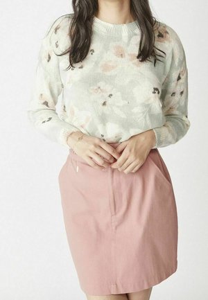 Woman wearing a white floral knit sweater tucked into a high-waisted pink skirt with pockets, hands clasped at waist.