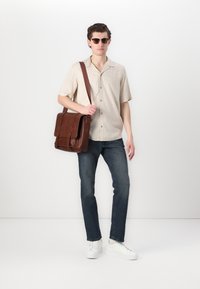 Bolso de hombro de cuero marrón con tapa, camiseta de manga corta beige con un patrón sutil, jeans azules y zapatillas blancas, sobre un fondo liso.