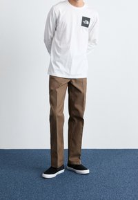 Weißes Langarmshirt mit einer schwarzen Logo-Tasche, kombiniert mit braunen, geraden Hosen und schwarzen Slippern mit weißen Sohlen.