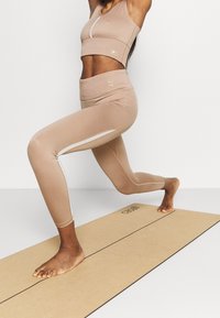 Completo fitness beige che presenta un reggiseno sportivo corto e leggings a vita alta con accenti bianchi, posato su un tappetino da yoga. Texture liscia.