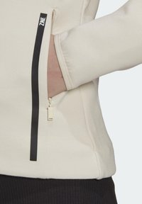 Béžová fleecová bunda s kapsou na zip a černým akcentem, hladkou texturou a vysokým límcem pro zvýšení tepla.