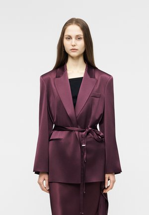 Vrouw met steil haar, gekleed in een satijnen pruimkleurige blazer met ceintuur over een zwarte top, staand tegen een effen lichtgrijze achtergrond.