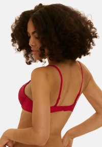 Frau mit lockigem Haar, die einen roten BH trägt und von hinten gezeigt wird, während sie ihn mit einer Hand vor einem schlichten weißen Hintergrund anpasst.