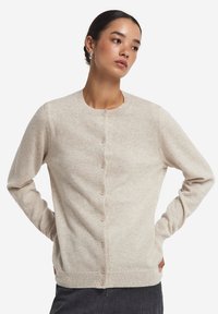 Beige stickad kofta med rund halsringning, långa ärmar och knappstängning fram. Materialet har en mjuk, teksturerad finish.