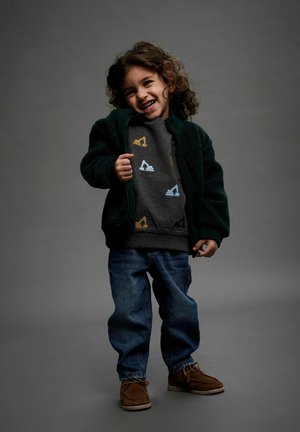 Kind trägt eine grüne Fleecejacke über einem grauen Sweatshirt mit bunten Baggerdesigns, ergänzt durch blaue Jeans und braune Schuhe.