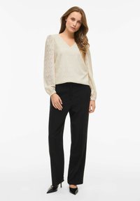 Beżowa bluzka o teksturze z dekoltem w kształcie V i długimi bufkami, zestawiona z dopasowanymi czarnymi spodniami i czarnymi szpilkami.
