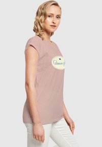 Lyserød t-shirt med korte ærmer og rund halsudskæring, prydet med et stort cremefarvet oval logo, der siger "Gilmore girls" i blå.