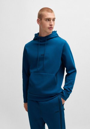 Sweat à capuche bleu en tissu doux, avec une poche frontale, des cordons de serrage et un logo. Associé à un pantalon assorti.