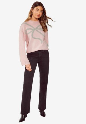 Rosa Pullover mit einem glitzernden grauen Schleifendetail, kombiniert mit schwarzen Jeans und spitzen schwarzen Ankle-Boots. Weicher Stoff, locker sitzend.