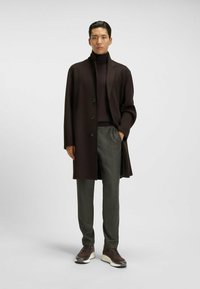 Mann in einem dunkelbraunen Mantel, dunkelbraunem Rollkragenpullover, grauen Hosen und braunen Turnschuhen vor einem einfarbigen Hintergrund stehend.