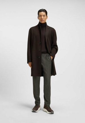Man wearing dark brown overcoat, dark brown turtleneck sweater, gray trousers, and brown sneakers standing against plain background.