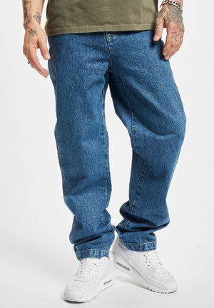 Vaqueros de mezclilla azul con un corte holgado, con costuras visibles, diseño de cinco bolsillos y dobladillo acampanado, acompañados de zapatillas deportivas blancas.