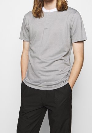 Jeune personne portant une chemise texturée gris clair à manches courtes sur un col blanc, avec un pantalon noir et les mains dans les poches.