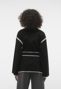 Schwarze Jacke mit langen Ärmeln und einem strukturierten Muster. Verfügt über einen dekorativen cremefarbenen Besatz und eine taillierte Passform. Glattes Material und lockere Passform.