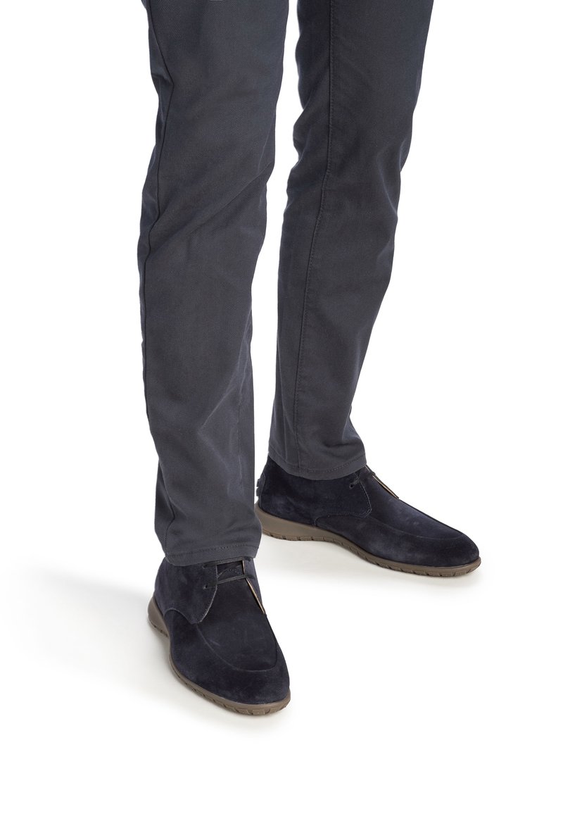 Personne portant un pantalon gris foncé et des chaussures à lacets en daim noir, debout sur un fond blanc.