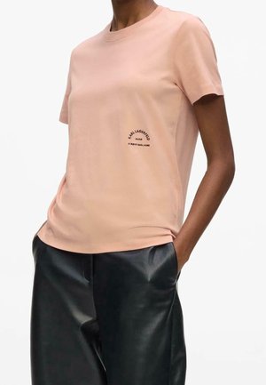 Person iført en lyserød kortærmet t-shirt med lille sort tekst på brystet kombineret med sorte læderbukser, hænderne i lommerne.