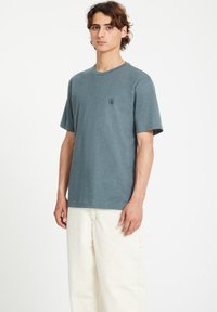 T-shirt en coton gris-bleu avec un col rond et des manches courtes, arborant un petit logo noir sur la poitrine gauche, porté avec un pantalon blanc.