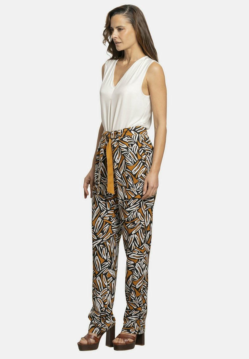Top blanco sin mangas combinado con pantalones de cintura alta de patrones en negro y marrón. Presenta un cinturón anudado y sandalias de plataforma gruesa.
