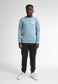 Felpa azzurra chiara con scollo a girocollo e toppa, abbinata a pantaloni cargo neri e sneakers bianchi. Presenta polsini e orlo a coste.