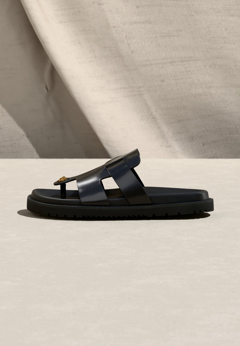 Black leather sandal with two wide straps and a toe post, flat sole, displayed on a light surface with a neutral background.