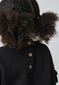 Felpa nera con texture a coste, caratterizzata da una patta e tre bottoni dorati come dettagli sul retro del collo. Capelli ricci visibili.