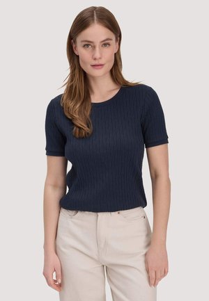 Frau mit langen braunen Haaren, die ein marineblau strukturiertes Kurzarmoberteil und hellbeige Hosen trägt und vor einem schlichten Hintergrund steht.