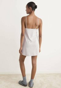 Femme avec un chignon, portant une robe nuisette blanche sans manches, des chaussettes grises et des pantoufles gris foncé, se tenant sur un tapis clair devant un mur sobre.