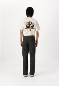 White t-shirt with back graphic depicting a black cow and figure against cacti and colorful sky; paired with brown trousers and gray shoes.
