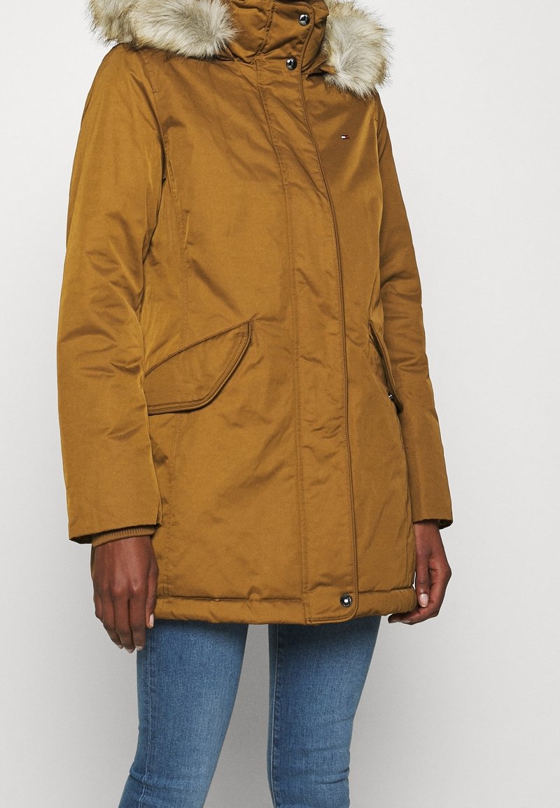 Manteau d'hiver marron avec capuche bordée de fourrure, porté avec un jean bleu, avec poches avant à rabat et fermeture par boutons-pression.