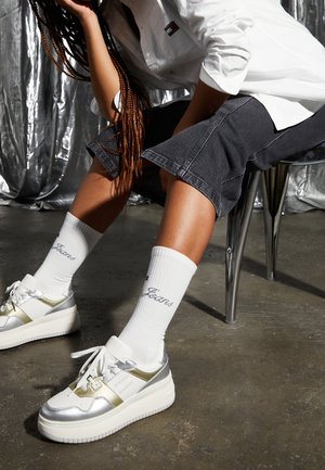 Białe sneakersy z srebrnymi akcentami i grubymi podeszwami, w połączeniu z białymi skarpetami z niebieskim wyhaftowanym logo oraz szarymi dżinsowymi szortami.