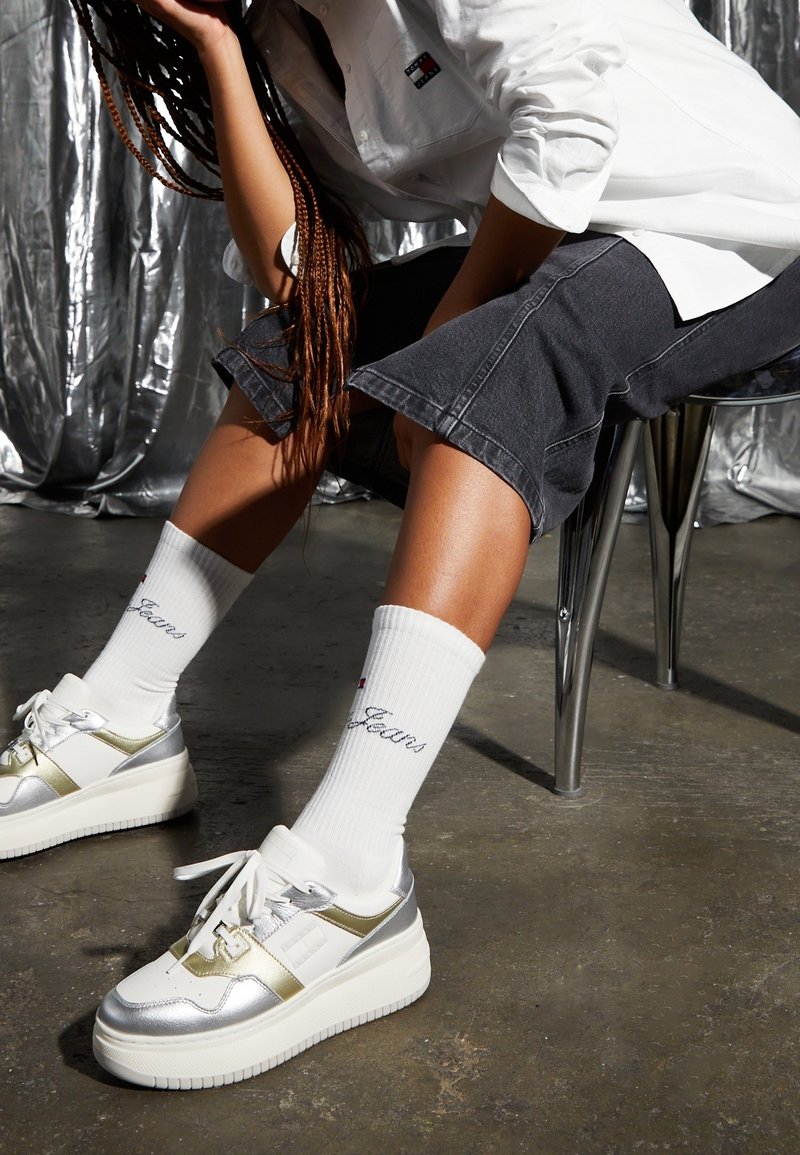 Białe sneakersy z srebrnymi akcentami i grubymi podeszwami, w połączeniu z białymi skarpetami z niebieskim wyhaftowanym logo oraz szarymi dżinsowymi szortami.