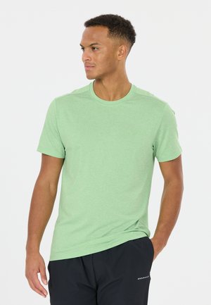 Mies, jolla on päällään vaaleanvihreä lyhythihainen T-paita ja mustat shortsit, seisoo käsi taskussa ja katsoo vasemmalle valkoista taustaa vasten.