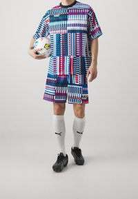 Conjunto deportivo multicolor con patrones geométricos en azul, rojo y blanco. Incluye pantalones cortos y una camiseta a juego, combinado con botas negras y calcetines blancos.
