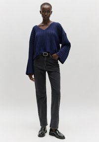 Marineblauer, gerippter Pullover mit V-Ausschnitt und weitem Ärmel, kombiniert mit hochtaillierten schwarzen Jeans und schwarzen, glänzenden Slippern. Glatte Textur.