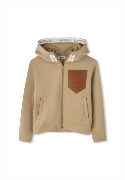 Beige Reißverschluss-Hoodie mit einem Lederpatch, das ein Logo zeigt, weicher Textur, Fronttaschen und einer kontrastierenden Kapuzenfutter mit Branding.