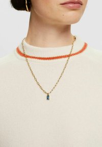 Collana in oro con maglie allungate, dotata di un pendente rettangolare in pietra azzurra. Indossata sopra un maglione crema chiaro con rifiniture arancioni.