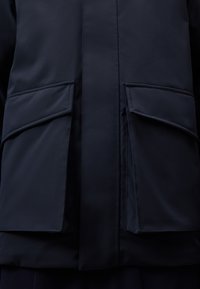 Marinblå jacka med en slät, matt yta, som har två stora framfickor. Designen inkluderar en rak skärning och minimala sömnadsdetaljer.